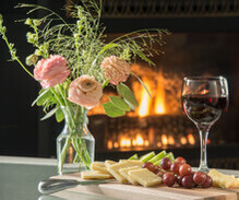 cheese plate, a glass of wine, and flowers on a table in front of a fireplace